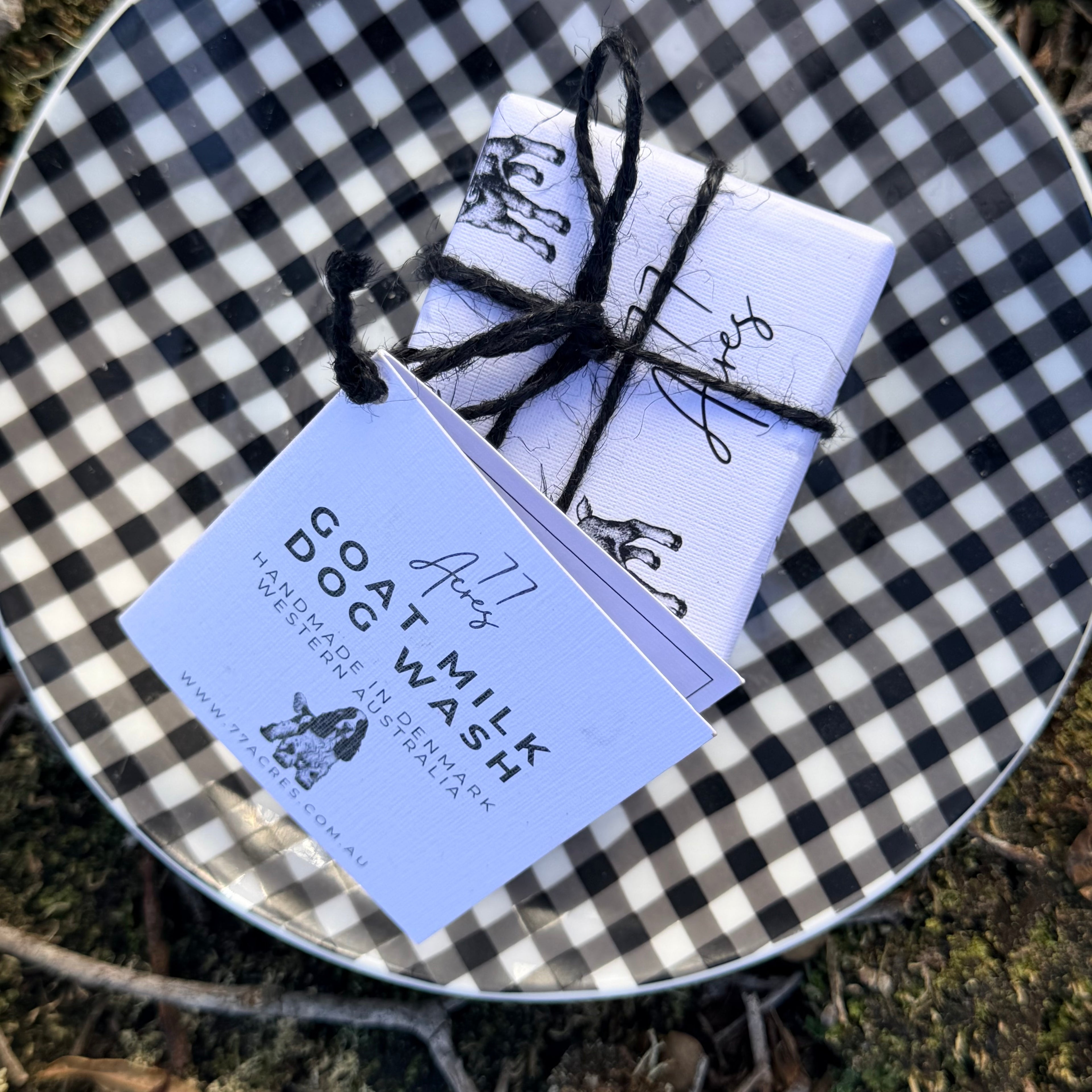 A packaged bar of soap wrapped in white paper with black goat illustrations, a label with an cocker spaniel dog and tied with black twine, placed on a black-and-white checkered plate outdoors on grass and wood.