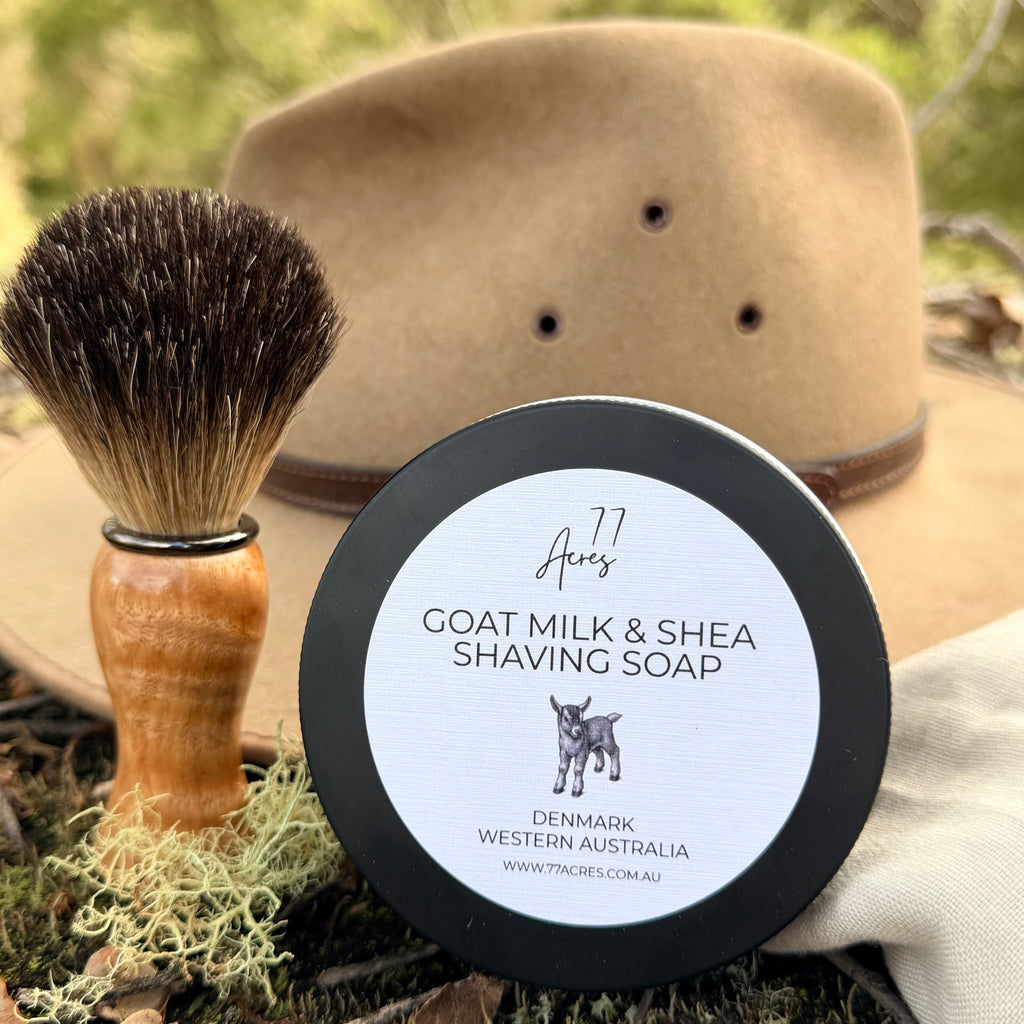 A round tin of 77 Acres  Goat Milk & Shea Shaving Soap in the foreground, with a shaving brush and a tan hat styled around it on a natural outdoor surface.