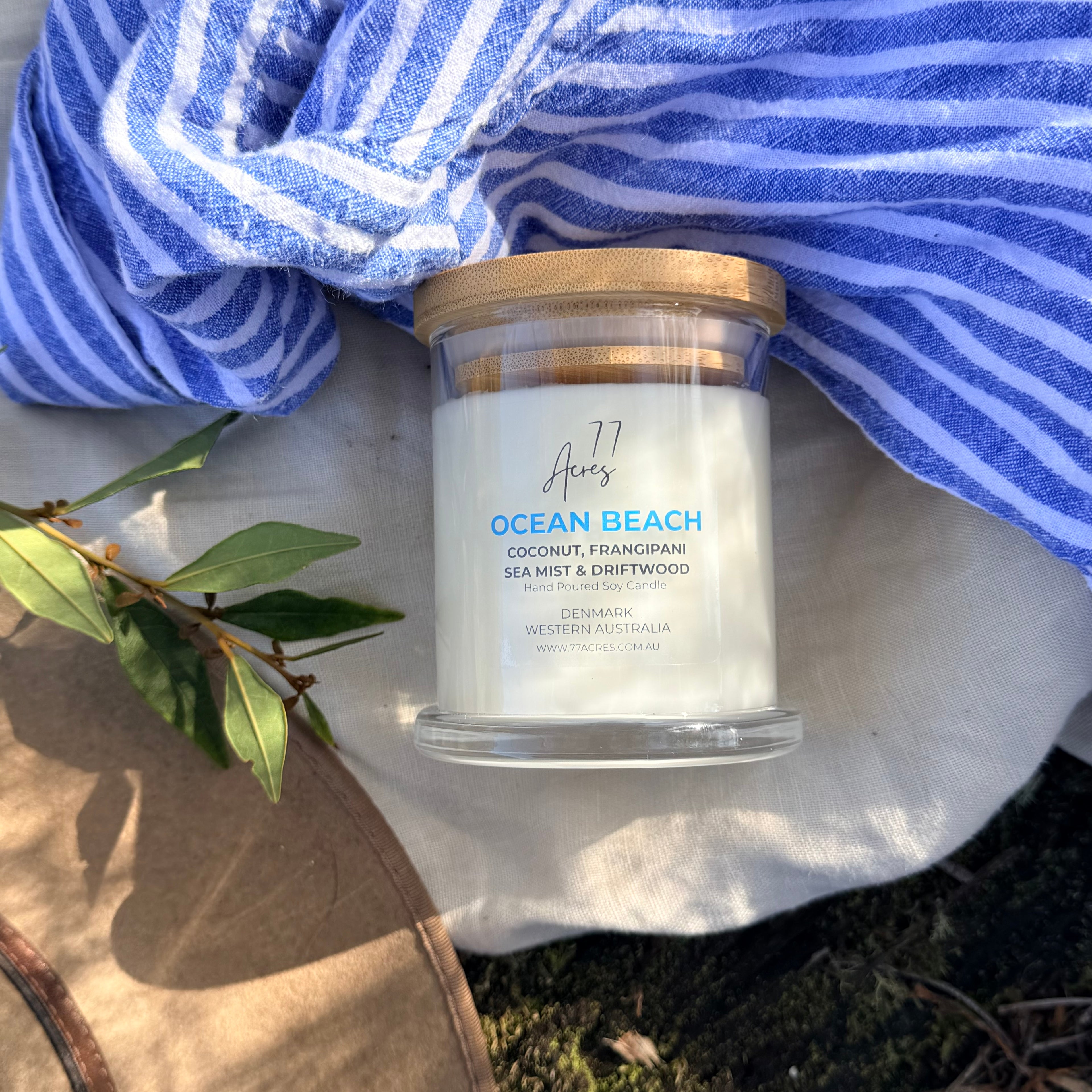 A glass jar candle labeled 'Ocean Beach' sits on a white cloth outdoors, styled with a blue-and-white striped towel, green leaves, and a wooden surface.
