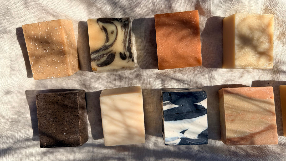 Nine rectangular bars of handmade soap in various colors and patterns, arranged in two columns on a light fabric background with dappled sunlight and shadows.