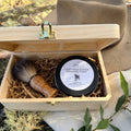 A black tim with white label displaying 77 Acres Goat Milk & Shea Shaving Soap. Presented in a wood box with a handmade wooden handle badger bristle shaving brush. Displayed in a bush setting with linen and wood. 