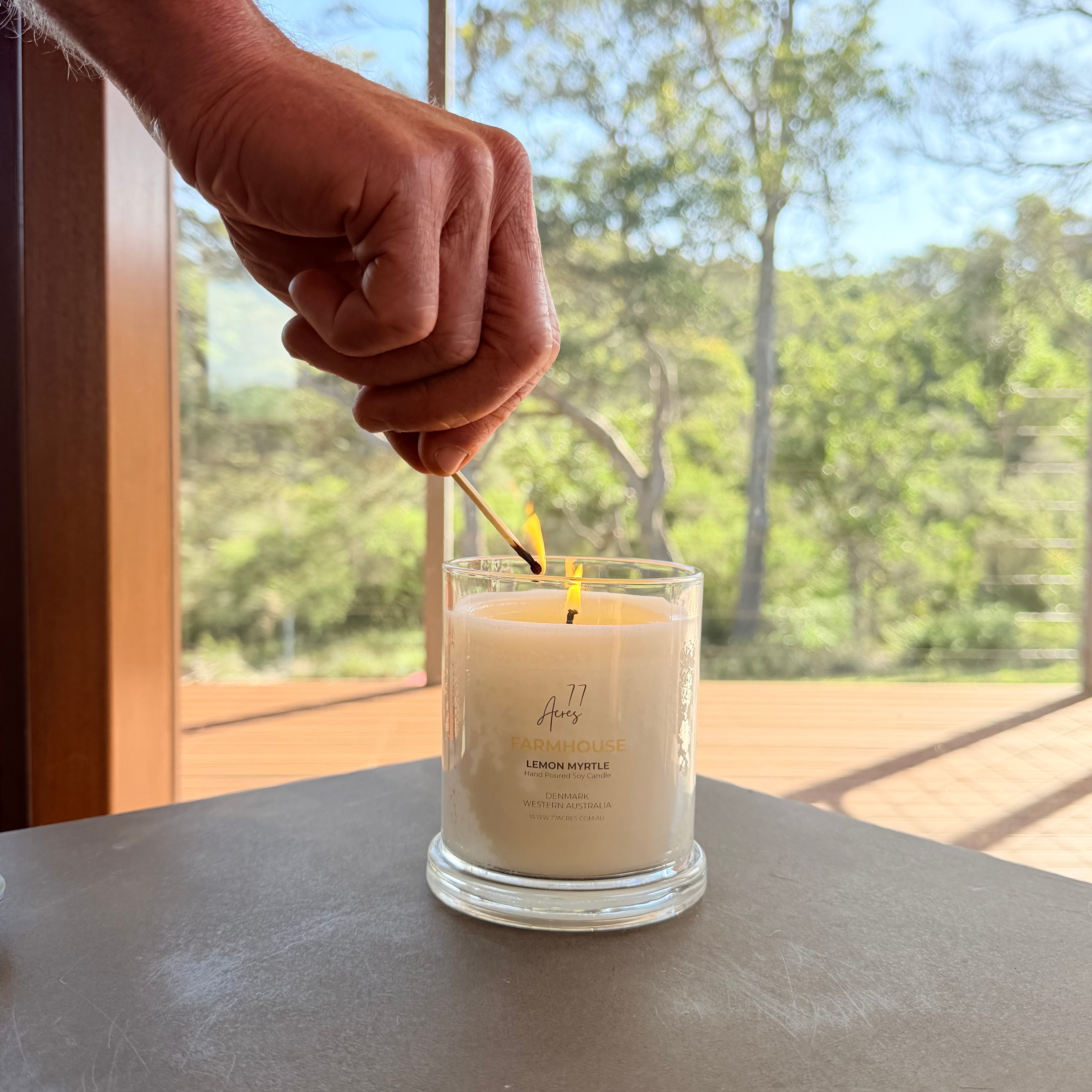 A hand lighting a candle in a 77 Acres clear glass jar with a match, set on a dark tabletop with an Australian bush background.