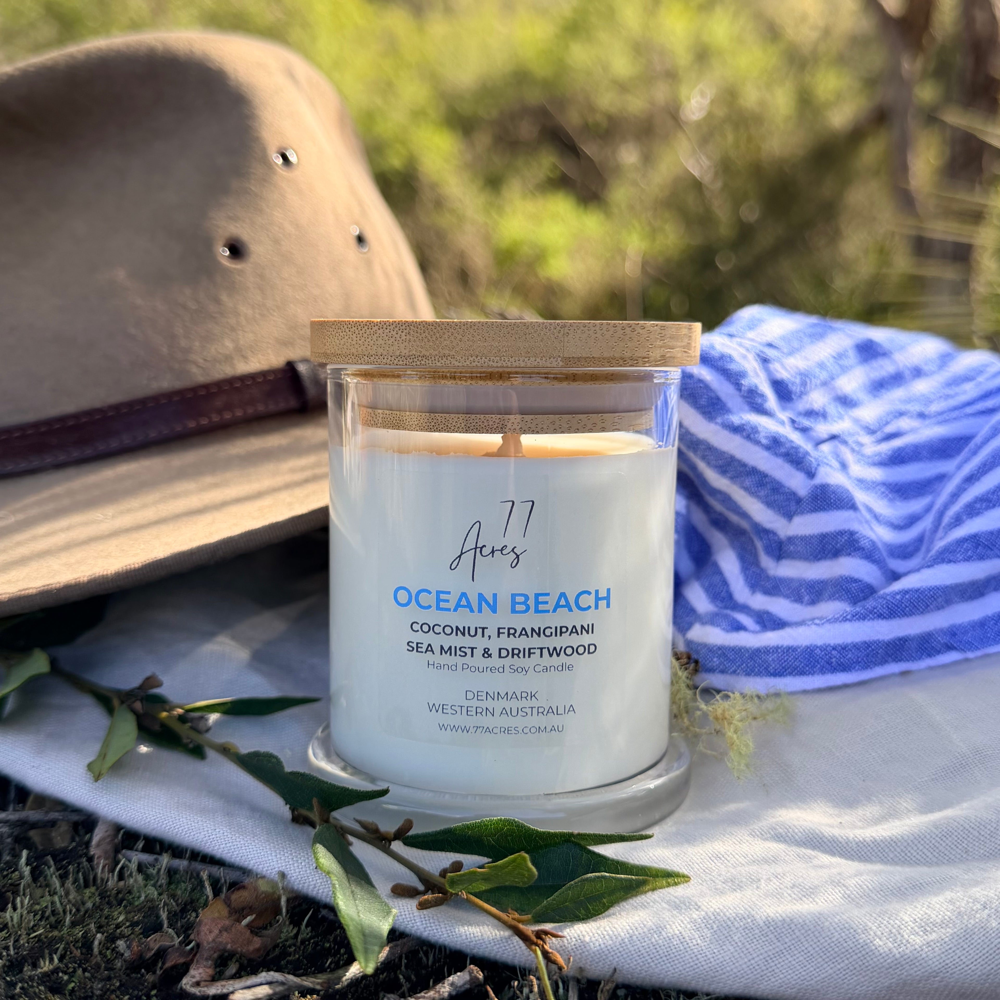 A jar candle labeled 'Ocean Beach' by 77 Acres Candle is styled outdoors on a white cloth with a blue striped towel, a tan hat, and green leaves. The candle is in a clear glass jar with a light wood lid and white wax.