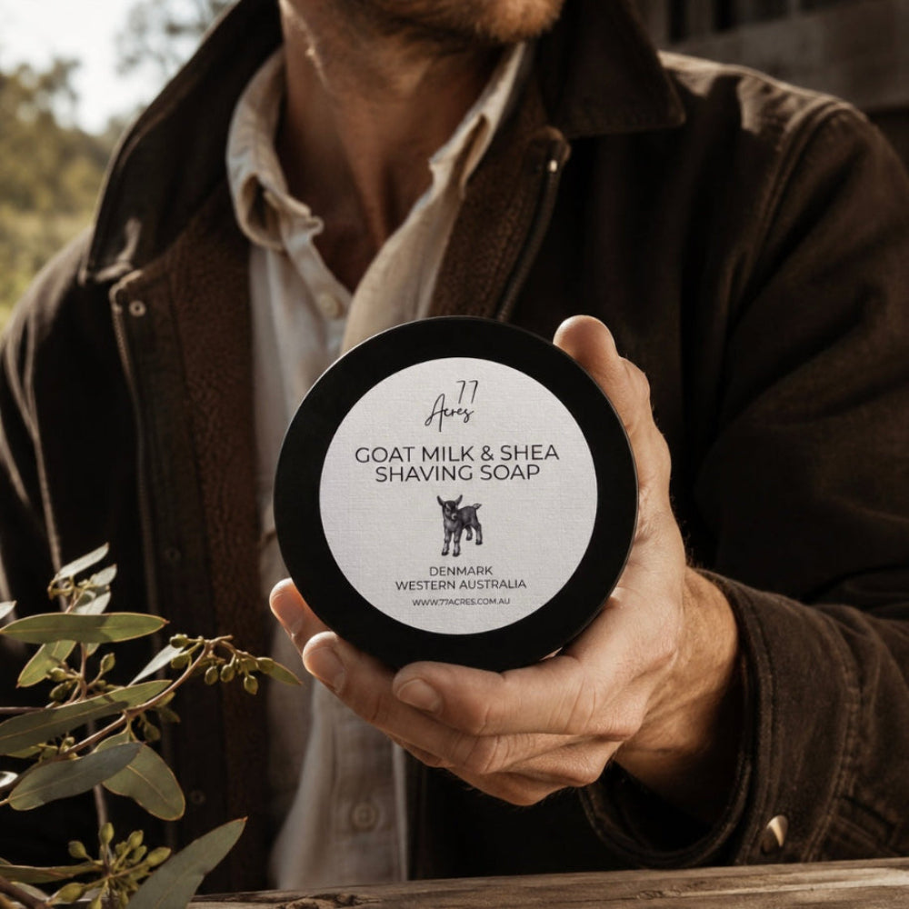 A man in a brown jacket holds a round black tin of Goat Milk Shaving Soap shaving. The label reads '77 Acres Goat Milk & Shea Shaving Soap' with a small goat logo and 'Denmark Western Australia'. 