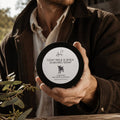 A man in a brown jacket holds a round black tin of Goat Milk Shaving Soap shaving. The label reads '77 Acres Goat Milk & Shea Shaving Soap' with a small goat logo and 'Denmark Western Australia'. 